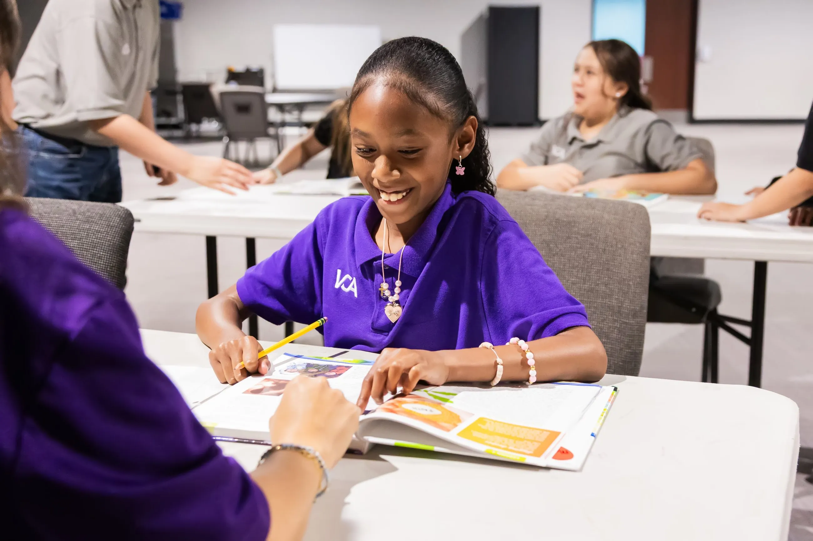 A student smiles while reading a workbook, guided by a teacher pointing at the page.