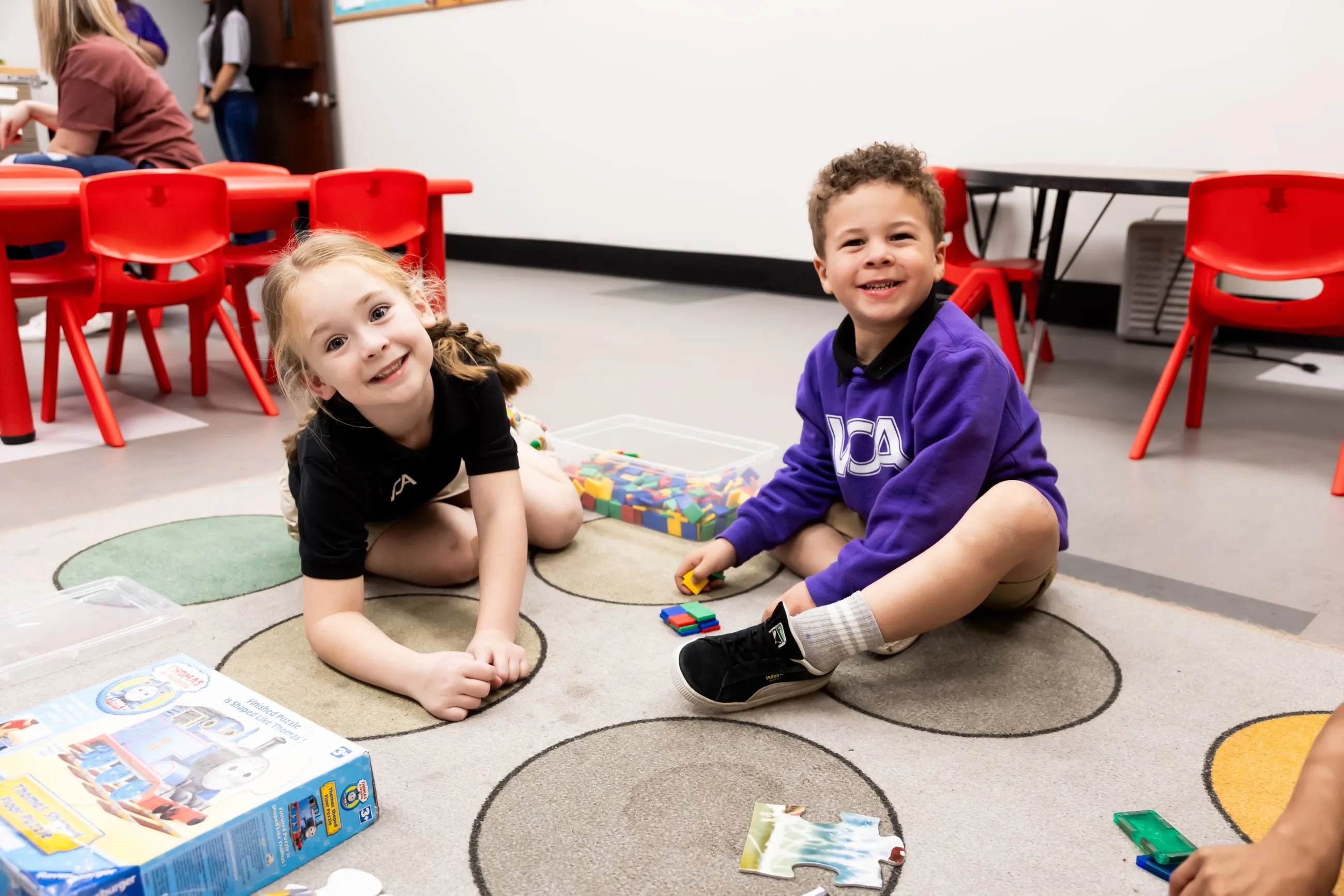 Two young students grin while playing with building blocks on the classroom rug.